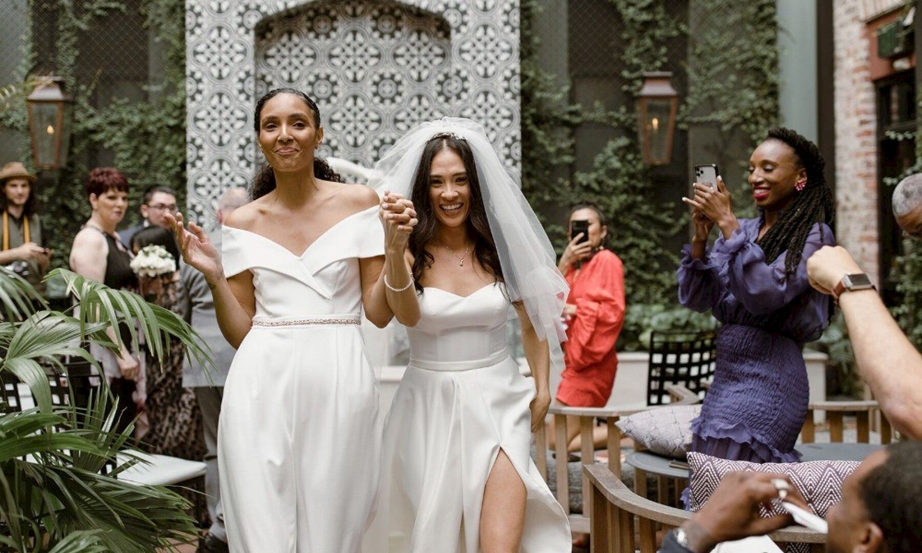 Two brides in white gowns walking arm in arm, smiling, as guests celebrate and take photos at an outdoor wedding setting.