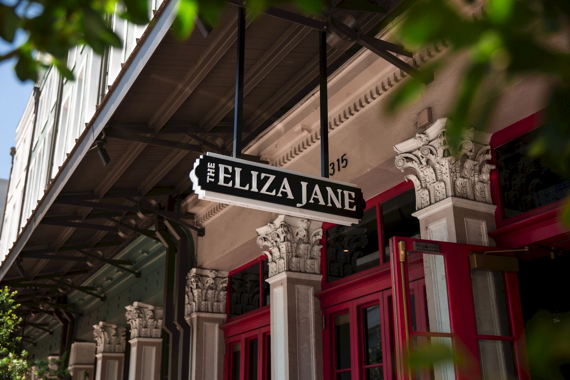 A storefront with ornate columns, a hanging sign that reads &ldquo;FELIZ A JANE&rdquo; and red doors, framed by greenery.