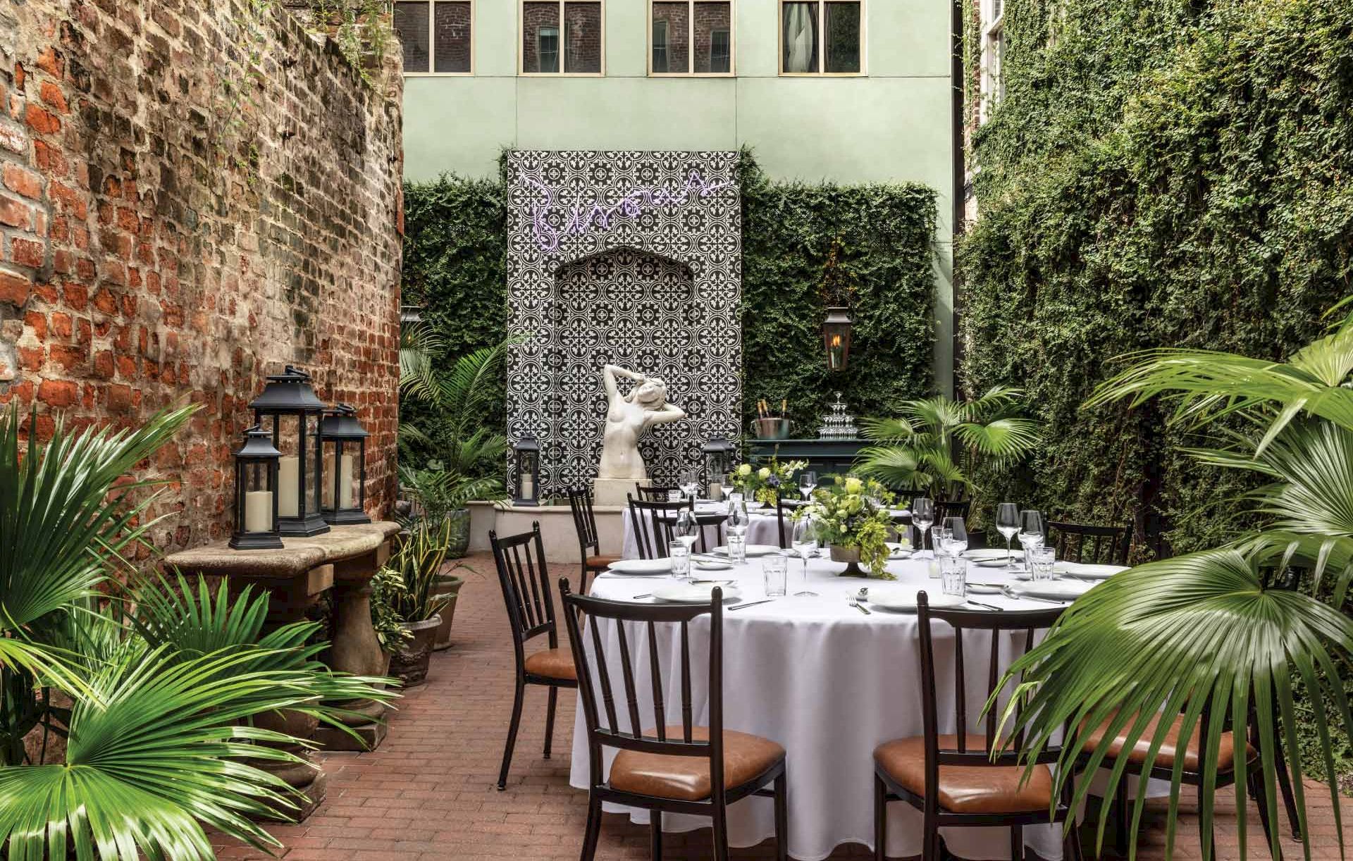 An intimate outdoor dining setup in a lush courtyard with a round table, white tablecloth, chairs, greenery, and a decorative tiled wall in the background.