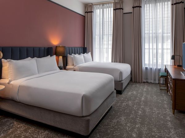 Two neatly made hotel beds in a modern room with a pink accent wall, lamps, curtains, and a dresser with a TV.