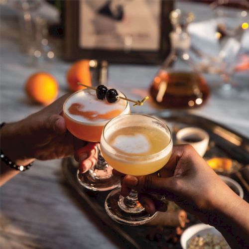 Two hands are toasting with cocktails in coupe glasses, garnished with cherries. A tray with drinks and fruit is in the background.