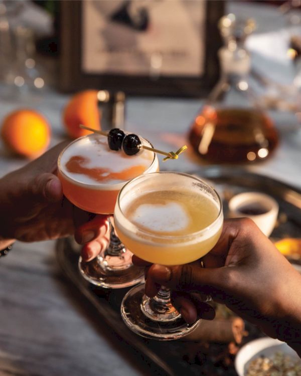 Two hands are toasting with cocktails in coupe glasses, garnished with cherries. A tray with drinks and fruit is in the background.