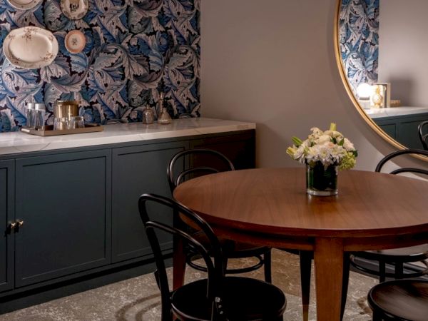 A round wooden table with dark chairs in a stylish dining nook, blue floral wallpaper, a sideboard with decor, and a mirror reflecting soft lighting.