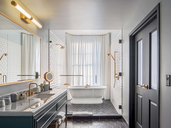 A modern, airy bathroom with a clawfoot tub, white tiles, brass fixtures, a double vanity, and a large mirror, light wood accents, and a dark door.