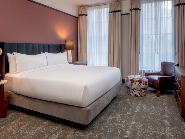 A clean hotel room with a large bed, nightstands, lamps, a seating area with a chair and ottoman, and a flat-screen TV on a dresser.