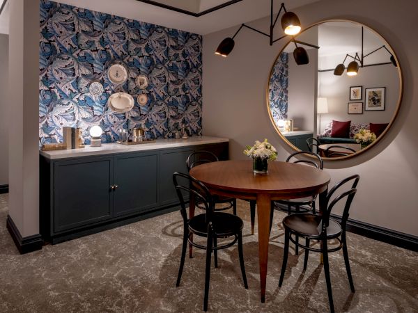A stylish dining nook with a round wood table, chairs, floral wallpaper, a buffet counter, and a large round mirror reflecting the room.