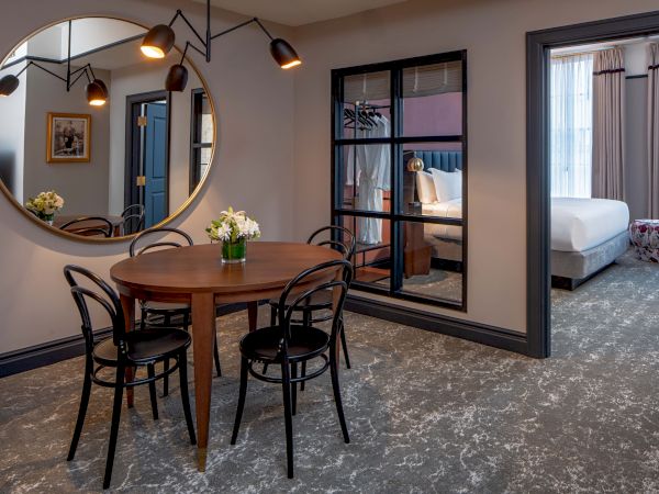 A modern dining area with a round table, black chairs, wall mirror, and open doorway showing a neatly made bedroom.
