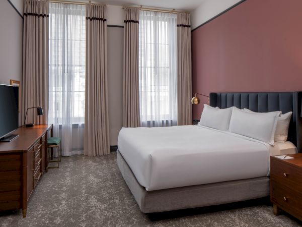 A modern hotel room with a king bed, nightstands, a TV on a wooden console, and large windows with beige curtains. (140 chars)