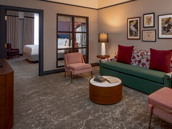 A cozy hotel suite: living area with a green sofa, pink chairs, round coffee table, framed art, and a lamp; bedroom visible through glass divider.