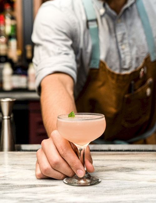 A bartender holds out a pink cocktail garnished with a mint leaf in a glass, in front of a bar setting with blurred bottles in the background.