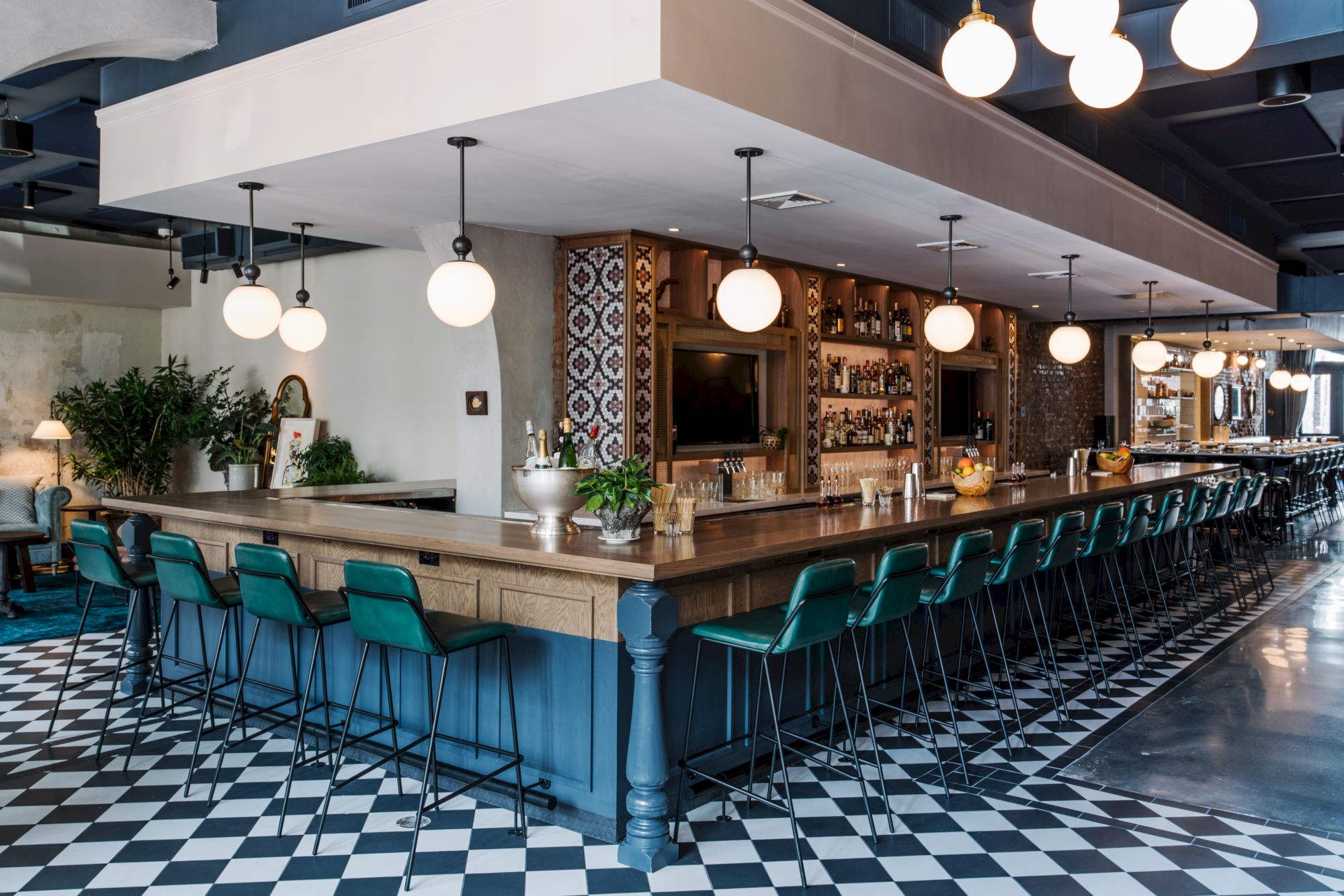 A long wooden bar with teal stools, tiled black-and-white floor, hanging globe lights, and shelves with bottles along a cozy, modern caf&eacute;/bar.