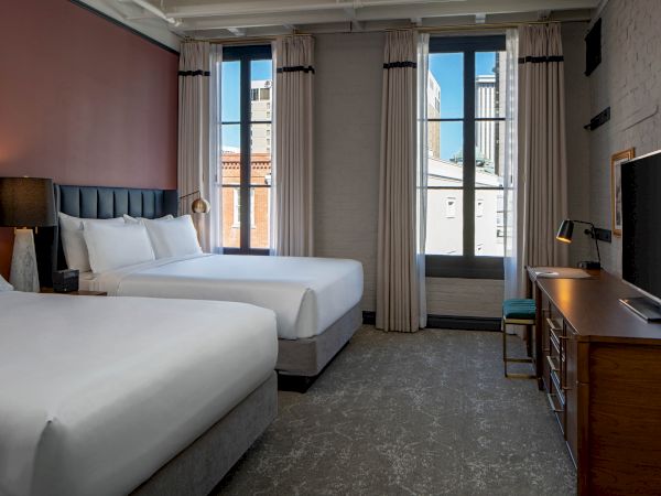 A hotel room with two queen beds, a desk and TV, large windows with beige curtains, and a neutral gray carpet.