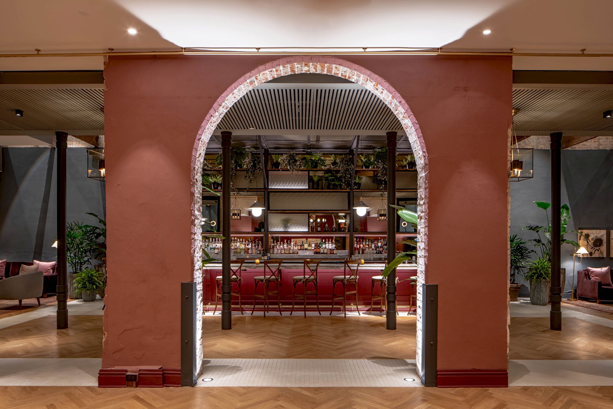 A warm, stylish restaurant interior with a red archway doorway opening into a bar area, wooden shelves, plants, and ambient lighting.
