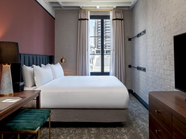 A modern hotel room with a king bed, white linens, a side desk and lamp, a bench, a dresser with a TV, and a window with curtains by city views.