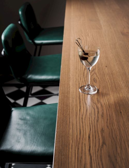 A dim, empty bar with teal leather chairs and a wooden counter, a cocktail glass garnished with a curl of zest perched at the edge.