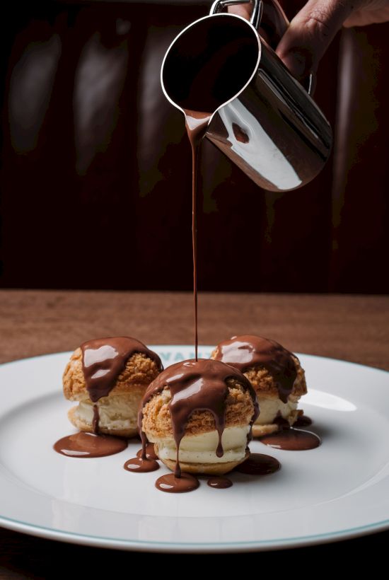Pouring glossy chocolate over creamy eclairs, silky caramel clouds drape the bite-sized indulgence over a white plate.