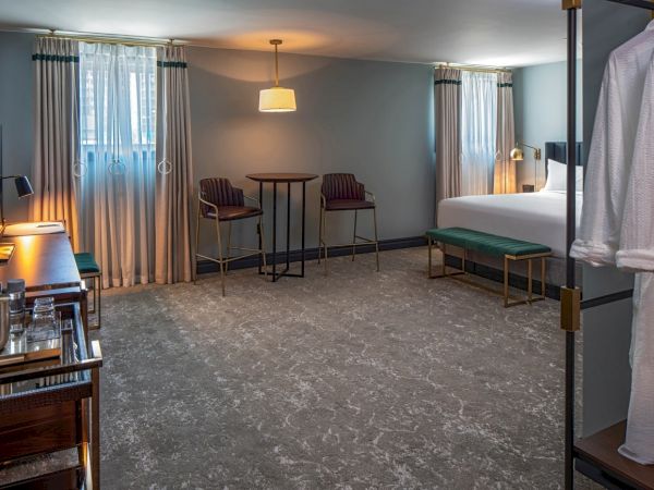 A hotel room with a large bed, seating area, desk, dresser, and robes in a spacious, softly lit, neutral-toned suite.