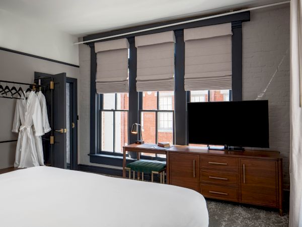 A hotel-style bedroom with a large bed, a dresser and TV, a windowed wall with blinds, and a small desk; an open closet area on the left.
