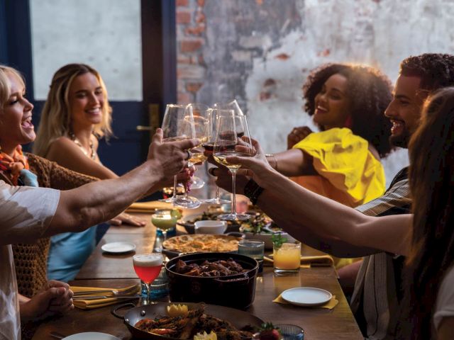 A group of people raising glasses in a toast at a cozy dinner gathering around a wooden table with food and candles.