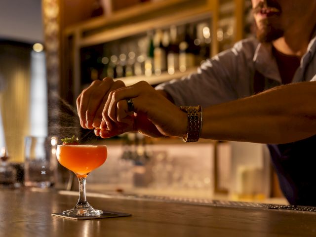 A bartender is garnishing a cocktail on a bar counter, with shelves of bottles in the background and a warm, inviting atmosphere.