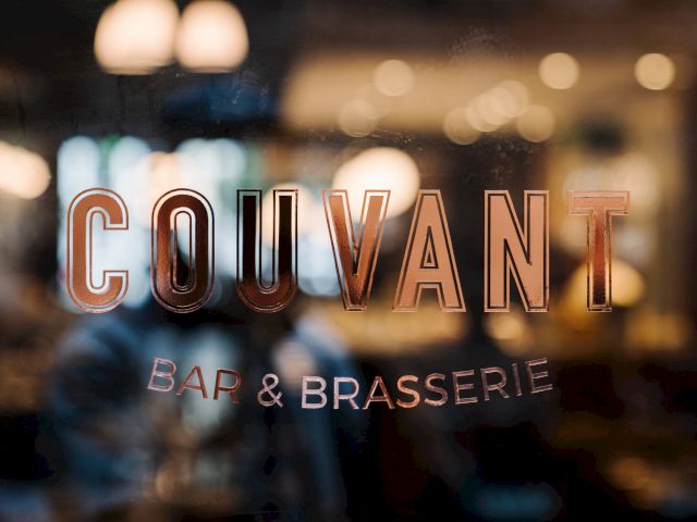 The image shows a blurred background with the words "COUVANT BAR & BRASSERIE" on a glass surface, suggesting a restaurant or bar setting.