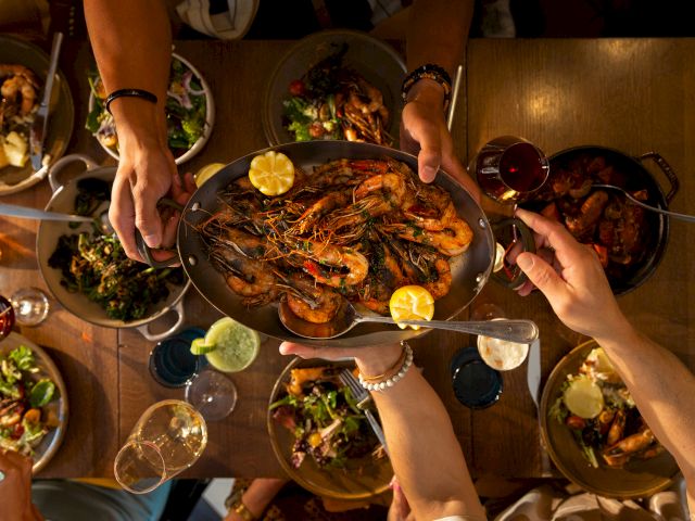 A group is sharing a meal, featuring a platter of shrimp with lemon, various dishes, and glasses of wine on the table.