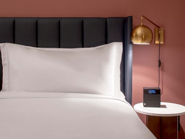 A neatly made bed with white sheets and pillows, a dark upholstered headboard, and a round bedside table with a clock and a gold wall lamp&mdash;soothing hotel vibe.