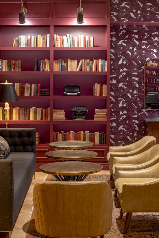 A stylish lounge with red walls, a bookshelves-filled nook, a gray sofa, and tan chairs facing a central wooden coffee table, plus a patterned wallpaper room in the background.