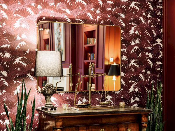 A room with red patterned wallpaper, a wooden table with a lamp, mirror, and plants; bookshelf reflected in the mirror.