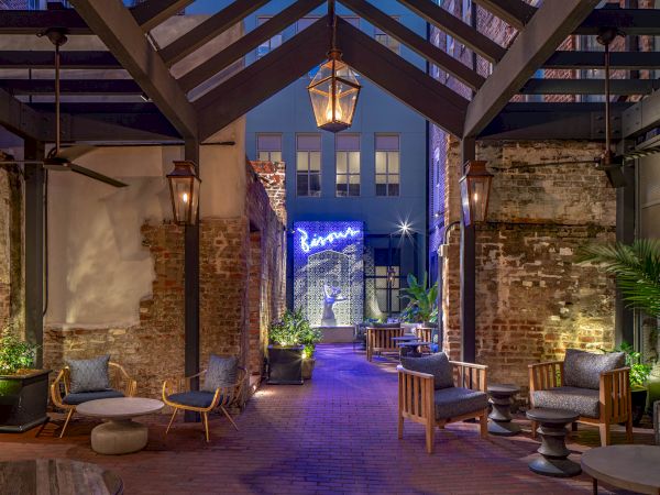 A cozy outdoor patio with seating, plants, and warm lighting. A neon sign and exposed brick walls add to the ambiance.
