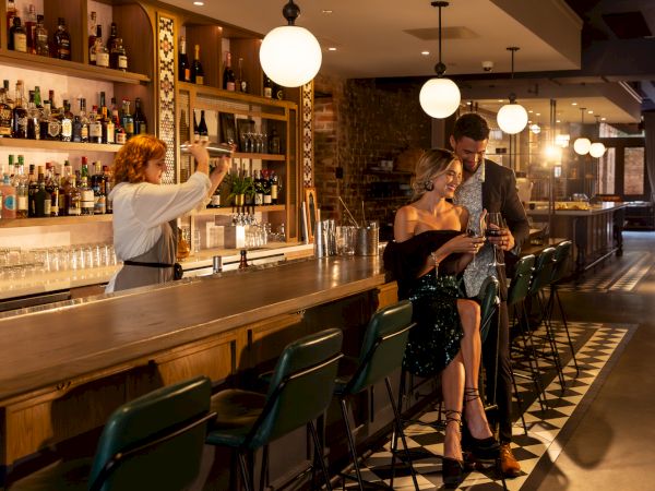 A cozy bar setting with a bartender preparing drinks and a couple standing at the counter, engaging with each other warmly.