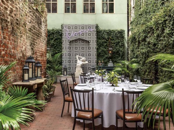 An outdoor dining setup with a round table, chairs, and lush greenery in a cozy courtyard with brick walls and decorative elements.