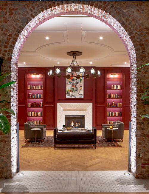 A cozy lounge area with an archway, plants, bookshelves, seating, and a fireplace, illuminated by warm lighting, creates an inviting atmosphere.