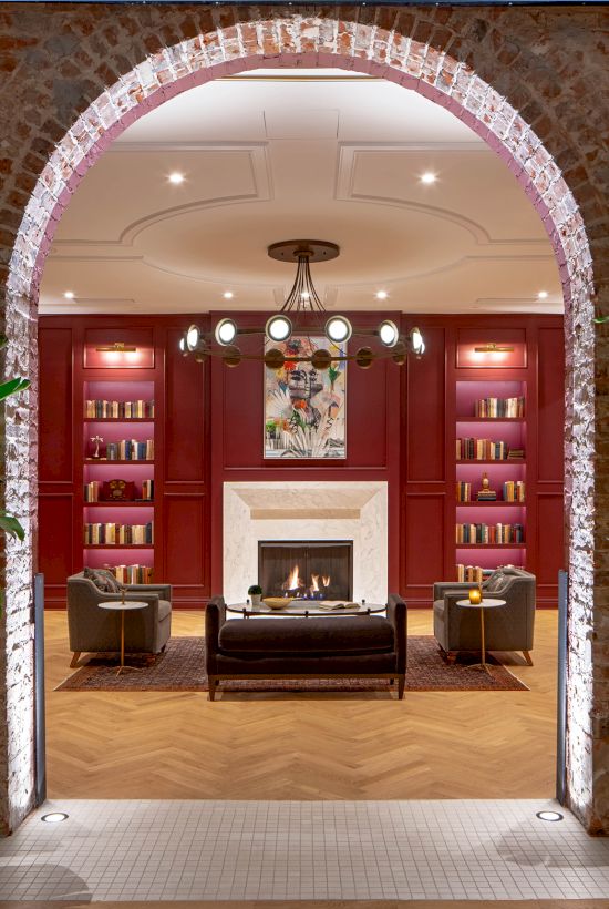 A cozy lounge area with an archway, plants, bookshelves, seating, and a fireplace, illuminated by warm lighting, creates an inviting atmosphere.