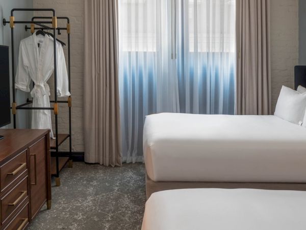 A hotel room with two beds, a desk, flat-screen TV, a clothes rack, desk lamp, bedside table, and a window with curtains.