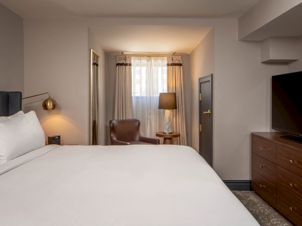 Hotel room scene: a large bed with white linens, nightstand lamps, seating area by the window, a dresser with a TV, and soft ambient lighting.