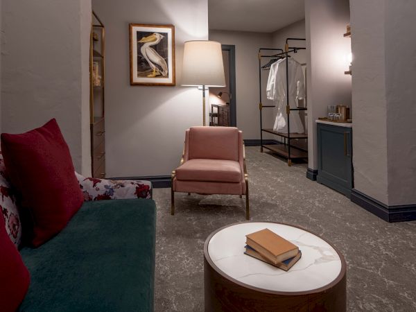 A cozy hotel suite: a teal sofa with red pillows, a pink chair, round coffee table with a book, soft lighting, and a wardrobe area in the background.