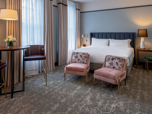 A luxurious hotel room with a king bed, soft lighting, two pink upholstered chairs, a small table, and a round desk area by tall windows.