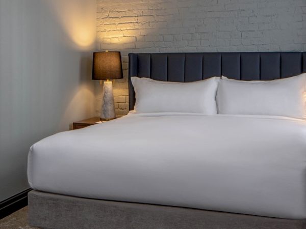 A modern hotel bedroom with a white bed, padded headboard, two lamps, exposed brick wall, nightstands, and a corner shelf unit in patterned wallpaper.