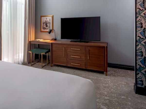 A hotel room with a bed in the foreground, a wooden dresser with a flat-screen TV, a small desk and chair, and a window with curtains.