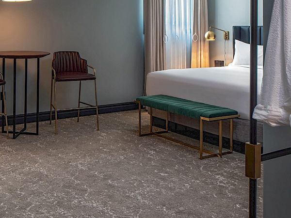 A modern hotel room with a bed, small round table and two chairs, a bench at the foot, a desk with a kettle, and soft lighting across a patterned carpet.