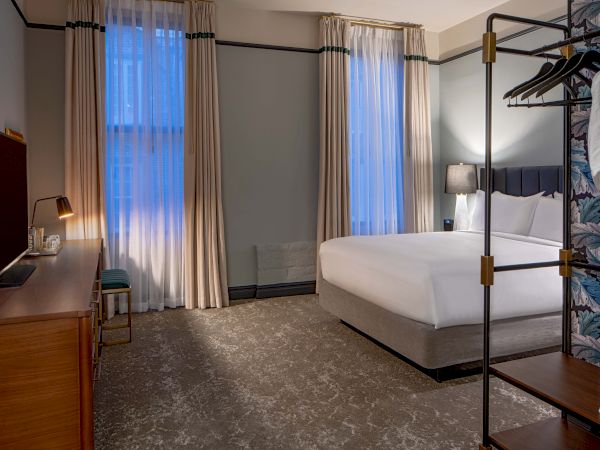 A hotel room with a large bed, bedside lamp, wooden desk, and floor-to-ceiling curtains letting in daylight, ending in a cozy setup.