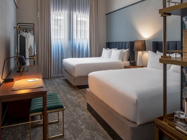 A hotel room with two double beds, a desk and chair, a wall art piece, a window with sheer curtains, and bedside lamps for lighting.
