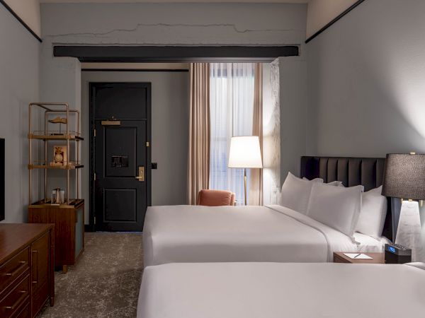A hotel room with two large beds, a nightstand lamp, a tv stand, a door, shelving unit, and a chair by a window with a floor lamp.