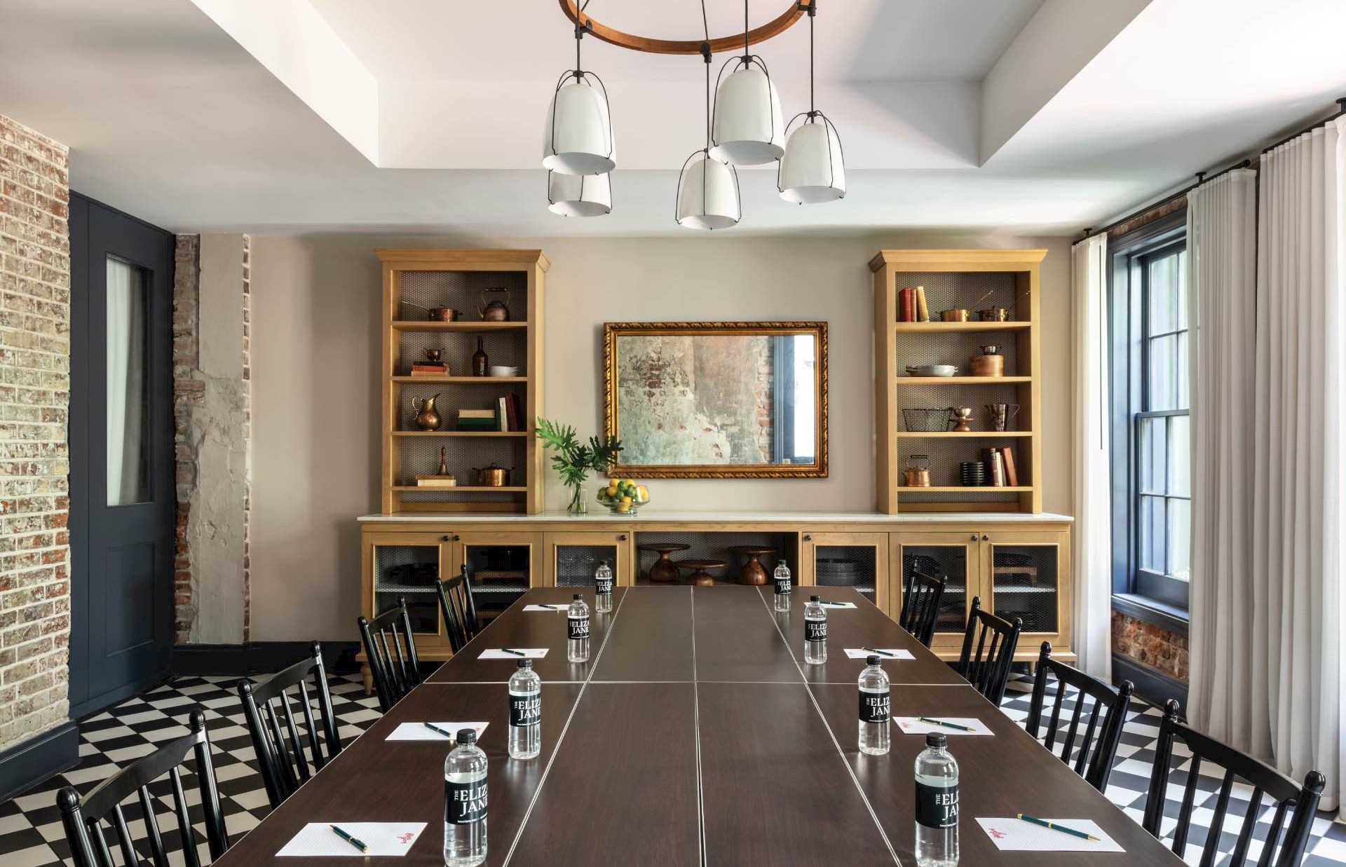 A modern meeting room with a long table, black chairs, shelves, a central mirror, brick wall, and large windows; inviting and organized.