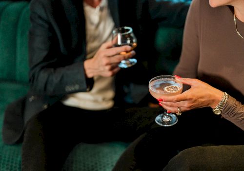 A couple sits on a green couch, each holding a stemmed glass of drinks, chatting and enjoying a social night out.