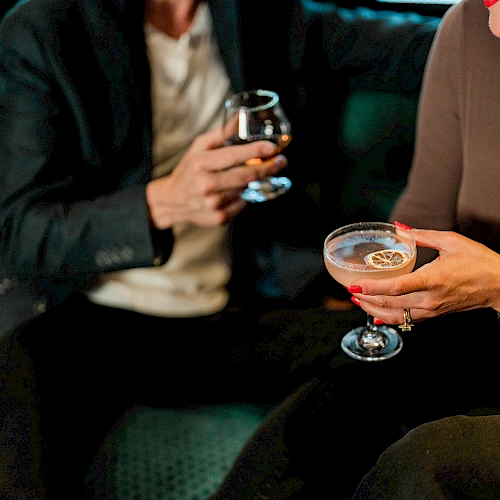 A couple sits on a green couch, each holding a stemmed glass of drinks, chatting and enjoying a social night out.