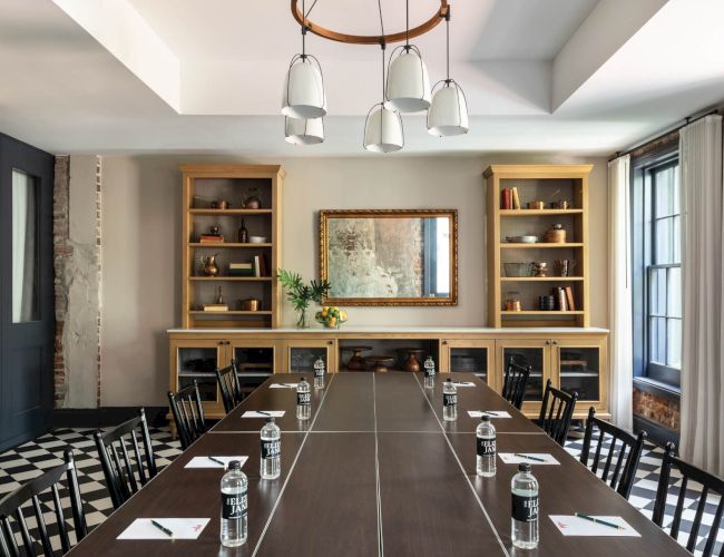A stylish conference room with a long table, black chairs, water glasses, notepads, and a built-in shelving unit flanking a central framed mirror; industrial yet warm.