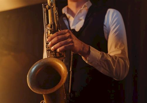 A person in a white shirt and dark vest plays a saxophone, close-up of hands on the keys and bell, warm stage lighting.