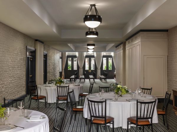 A stylish dining room with round tables, white tablecloths, black chairs, potted greenery, and soft hanging lights in a brick-walled, elegant venue.